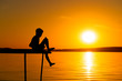 © Vadim - Silhouette of a happy boy sitting on the end of a bridge over the river at sunset. A small boy sits alone on a wooden bridge on the background of a river in the evening.