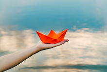 Nice Red Paper Boat On The Hand Of A Young Woman Above The Water In The River Background. Woman Holding Origami Boat Near River. Close-up