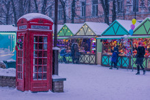 Christmas Holidays Concept Of Red Classic English Phone Booth, Season Winter Fair With People Background In Snowing Weather 