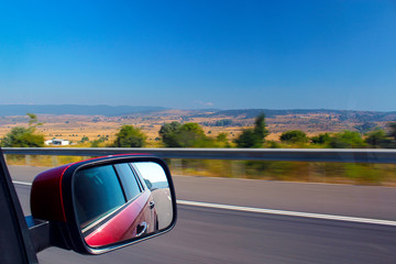 Red SUV driving at speed on the road. In the rear view mirror displays the roadbed with the markup and the car itself. View of the mountain landscape from the car window.