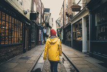 A Rear View Of A Girl In A Yellow Coat Walking Along The Historic Street Known As The Shambles In York, UK Which Is A Popular Tourist Destination And Medieval Landmark In This Ancient City