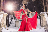 beautiful young girl dancing belly dance in red dress in winter in a park on the snow.