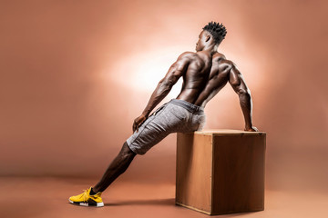 Wall Mural - Muscular African American Black athletic man sitting back to camera  on the edge of a box in studio in his blue underwear with dramatic lighting with a brown background   