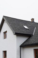 Part of white house with new wooden eaves, plastic rain gutter, window and chimney in grey shingles roof and brown window in facade  with copy space on sky.