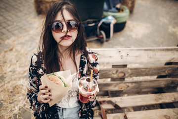 Wall Mural - stylish hipster woman holding juicy burger and lemonade. boho girl with hamburger and drink smiling at street food festival. summertime. summer vacation travel. space for text