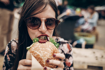 Wall Mural - stylish hipster woman eating juicy burger. boho girl biting yummy cheeseburger, smiling at street food festival. summertime. summer vacation travel picnic. space for text