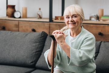 Wall Mural - cheerful senior woman sitting on sofa and holding walking stick