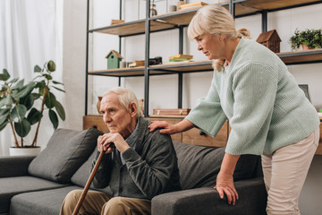 Wall Mural - senior wife standing near retired husband sitting on sofa