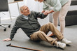old woman helping to stand up husband who falled down on floor near walking stick
