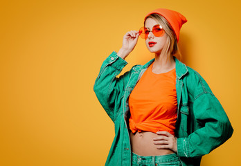 Wall Mural - Young style girl in jeans clothes with orange glasses on yellow background. Clothes in 1980s style