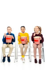 cheerful kids sitting on chairs, holding striped buckets and eating popcorn isolated on white