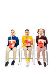  smiling children sitting on chairs, holding striped buckets and eating popcorn isolated on white