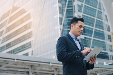 young asian investor business man in modern suit standing and working about finance on digital mobile tablet in office building, smart technology, investment, stock market and global business concept