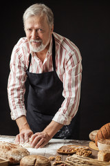 Mature male baker with grey beard, looking at camera, preparing bread dough on wooden table in a bakery with a variety of bakery products on foreground. Preparation of Easter bread.