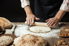 Chef Or Baker, Dressed In Black Apron, Preparing A Portion Of Fresh Dough In Rural Bakery, Kneading The Pastry Surrounded By Rustic Organic Loaf Of Bread.