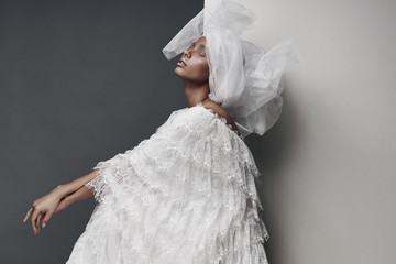 Wall Mural - Studio portrait of woman with white make up, in white lace dress, white veil head tie and big earring. Grey and white background