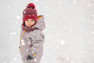 Wall Mural - Portrait of a cute baby dressed in a gray jacket and a red hat that walks through the snow covered park  enjoying first snow blowing