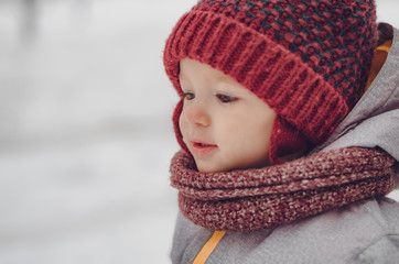 Canvas Print - Portrait of a cute baby dressed in a gray jacket and a red hat that walks through the snow covered snow park. She smiles one in the photo during the snowfall