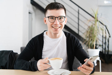 Indoor shot of young smiling man sitting in coffee shop. Handsome caucasian guy at cafe using smart phone.