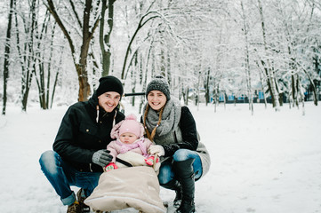  Happy family walks in the park. full length. Mom, dad, daughter stand outdoors on the background winter. Close up. Portrait baby in children's sled. Infant.