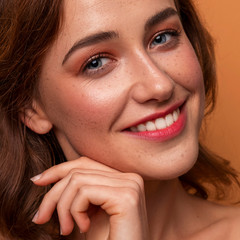 Cute caucasian brunette woman with curly hairstyle on orange background. Fresh clean skin with evening make up and freckles. Hands near her face. Close up portrait