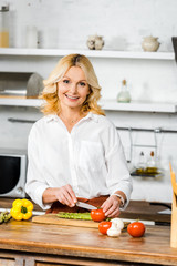 Wall Mural - smiling attractive middle aged woman cutting vegetables in kitchen and looking at camera