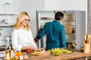 mature wife cutting vegetables and husband looking at fridge in kitchen