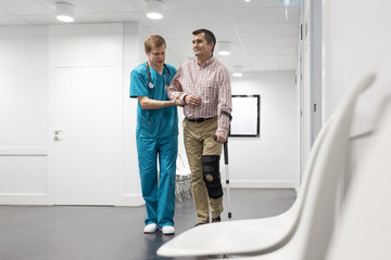 Nurse assisting patient with injured leg in walking at hospital corridor