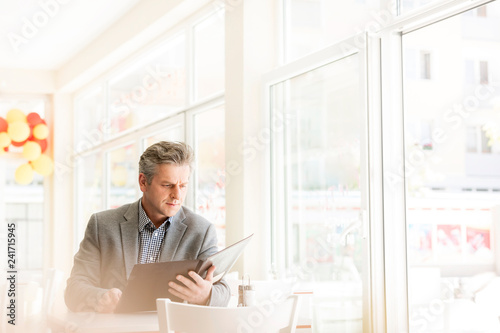 Fotografía Mature customer reading menu while sitting at table by window in restaurant