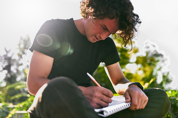 Poster - Outdoors image of handsome male student with curly hair, writing some notices on paper, wearing black t-shirt sitting on the street. Freelancer businessman makes plan for orders