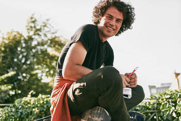 Poster - Side view of man student with curly hair, smiling and wearing black t-shirt sitting on the street and preparing for exams. Freelancer businessman planning the day sitting on the city street