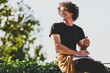 © iuricazac - Successful smiling male student with curly hair, smiling and wearing black t-shirt sitting on the street and preparing for exams. Freelancer businessman planning the day sitting on the city street