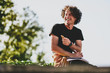 © iuricazac - Confident happy guy with curly hair, smiling and wearing black t-shirt sitting on the street and preparing for exams. Freelancer businessman planning the day sitting on the city street