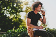 © iuricazac - Side view portrait of good-looking man student with curly hair, wearing black t-shirt, eating snack and preparing for exam. Freelancer businessman planning the day sitting on the city street