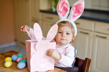 Cute little toddler girl wearing Easter bunny ears playing with colored pastel eggs. Happy baby child unpacking gifts. Adorable healthy smiling kid in pink clothes enjoying family holiday