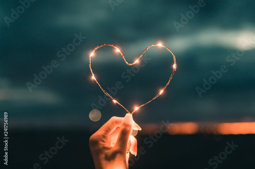 Fotomural Simbolo del cuore fatto da una striscia di luci led tenuto da una mano di una ragazza di fronte al cielo sfuocato