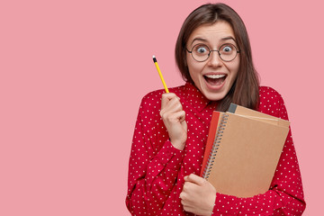 Wall Mural - Cute young woman with joyful expression carries pencil and notepad, opens mouth widely, makes notes in notebook, stands over pink background with blank space on right side for your promotion.