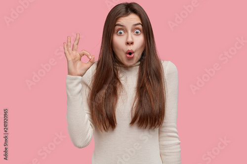 Fotografie, Obraz Studio portrait of shocked amazed woman makes okay gesture with suprised expression, stands over pink background, dressed in white casual jumper, amazed by something