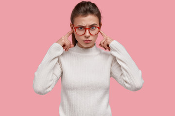 Wall Mural - Dissatisfied young woman plugs ears with index fingers, has irritated facial expression, avoids negative sound, annoyed with loud music, dressed in white clothes, isolated over pink background