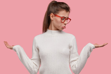 Wall Mural - Horizontal view of upset hesitant indifferent woman purses lip, raises palms, has pony tail, indecisive expression, wears spectacles, holds two things in hands, isolated over pink background.