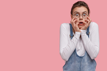 Wall Mural - Human facial expressions and emotions concept. Woman in despair and shock, stands in stupor against pink background, touches face, has scared look, wears round spectacles and denim overalls.