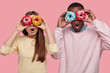 Horizontal shot of surprised dark skinned man covers eyes with delicious sparkling doughnuts, stands near her girlfriend, spend free time together, like eating dessert, isolated over pink background