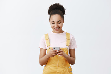 Wall Mural - Cute carefree girl making schedule for tomorrow with new app. Portrait of charming urban female with dark skin in yellow overalls, holding smartphone, smiling at screen while typing message