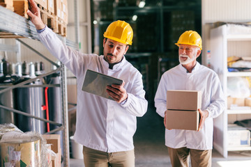 Young male Caucasian worker using tablet while standing in warehouse. In background other worker carrying boxes.