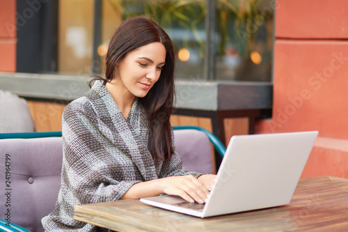 Foto Shot of female freelancer creats publication for content blog, uses modern gadget, keyboards on laptop, poses on sofa in cafeteria
