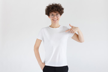 Wall Mural - Young woman pointing with index finger at blank white t-shirt with empty space for your advertising text or image, standing isolated on gray background