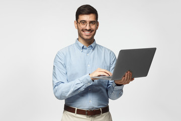 Wall Mural - Confident young handsome business man in blue shirt holding laptop and smiling at camera, isolated on gray background