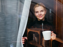 Adorable Woman Looking Out Through An Window And Holding A Cup Of Coffee Or Tea. Beautiful And Thoughtful Blonde With Curly Hair Standing Behind The Window.