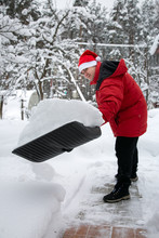 Man In Red Down Jacket And Red Hat Of Santa Claus Clears Snow In Backyard. Clears Snowdrifts On Path To Home. In Snowfall. Man Remove Snow With Big Plastic Shovel With Wooden Handle