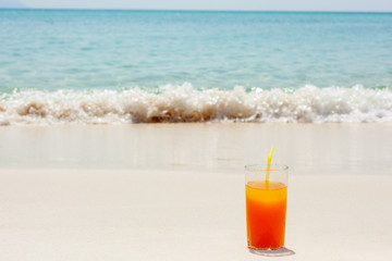 Wall Mural - Glass cup with orange juice with a straw on the beach on white sand against a blue sea